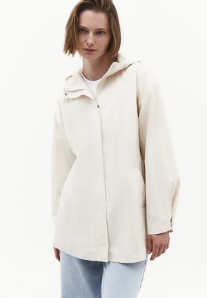 Mujer con chaqueta holgada con capucha de color beige claro con cremallera y bolsillos laterales combinada con vaqueros azul claro sobre un fondo liso.