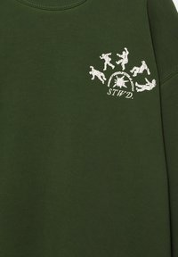 Sötétzöld ing, fehér grafikával, amely öt kis táncoló figurát ábrázol egy napkitörés körül, és az "STWD" betűk alatt.