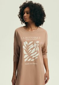 Beżowa, oversize koszula z rękawami 3/4. Posiada biały wzór roślinny oraz napis "LA BOTANIQUE" powyżej i "Beaute des Plantes" poniżej.