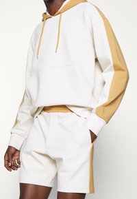 Vit hoodie med kamelfärgade detaljer och känguruficka, tillsammans med matchande vita shorts. Tyget verkar mjukt med en avslappnad passform och dragsko-detaljer.