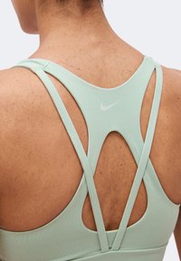 Κοντινό πλάνο του πάνω μέρους της πλάτης ενός ατόμου που φοράει ένα ανοιχτό πράσινο αθλητικό σουτιέν Nike με σταυρωτούς ιμάντες και ένα μικρό λογότυπο Nike στη μέση της πλάτης.
