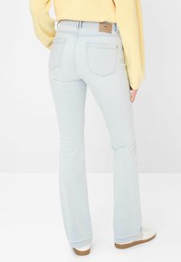 Persona con jeans azzurri a zampa d'elefante e una maglietta gialla a maniche lunghe, in piedi, vista posteriore con scarpe da ginnastica bianche.