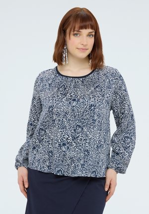 Vrouw met schouderlang rood haar draagt een blauw-wit blouse met bloemenpatroon en een marineblauwe rok, poserend tegen een effen achtergrond.