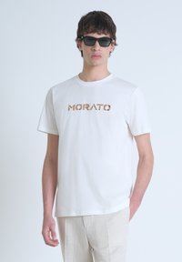 Camiseta blanca de algodón con un logo "MORATO" texturizado en marrón; modelo lleva gafas de sol negras y pantalones beige claros. Corte casual.