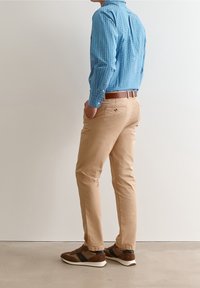 Camicia a maniche lunghe a scacchi blu, pantaloni beige e sneakers marroni con dettagli neri, mostrato di lato su uno sfondo semplice.