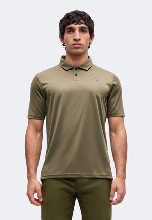 Hombre con una camiseta polo de manga corta verde oliva y pantalones a juego, de pie frente a un fondo claro y liso.