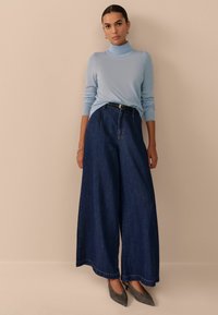 Lichtblauwe coltrui, donkere wijde denim broek, zwarte riem, grijze hakken. Gladde textuur, relaxte pasvorm, contrasterende kleuren.