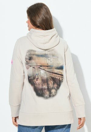 Kvinde med langt brunt hår iført beige hættetrøje med print af en sti til stranden med måger og solnedgang på ryggen.