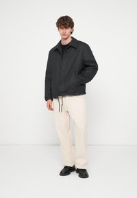 Sötétszürke kabát gallérral, gombos lezárással és húzózsinóros szegéllyel. Krémszínű nadrággal és fekete cipővel párosítva. Simaszövetű.