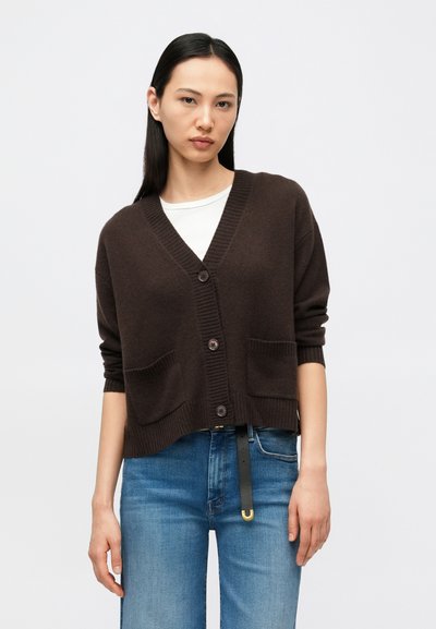 Brown knitted cardigan with a V-neck, button closure, and two front pockets. Textured fabric and cropped fit. Worn over a white shirt.