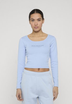 Vrouw met donker haar draagt lichtblauwe cropped top met lange mouwen en bijpassende broek, staand tegen een effen witte achtergrond.