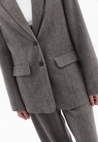 Harmaa herringbone-kuosisakkeli, jossa on kaksi ruskeaa nappia, kapeat läpät ja sivutaskut, valmistettu tekstuuripintaisesta kankaan kudoksesta.