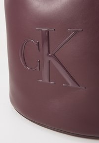 Borsa in pelle bordeaux con un logo "ck" in rilievo e lucido. Texture liscia con forma arrotondata e senza ulteriori accessori.