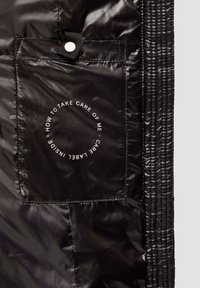 Schwarzer, glänzender Pufferjackenstoff mit einer quadratischen Tasche. Sichtbares Etikett mit weißem Text "WIE MAN FÜR MICH SORGT" in einem Kreis.