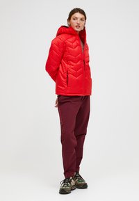 Rote Pufferjacke mit Chevron-Steppung, Kapuze und Reißverschluss vorne. Kombiniert mit bordeauxfarbenen Hosen mit seitlichen Taschen und einem orangefarbenen Logo. Braune Sneakers.