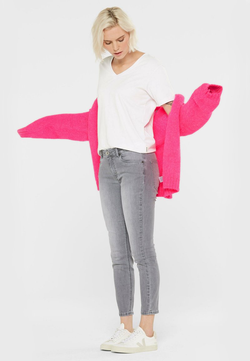 Fuchsia übergroßer Cardigan, der über einem weißen V-Ausschnitt-T-Shirt getragen wird, kombiniert mit grauen engen Jeans und weißen Sneakern für einen lässigen Look.