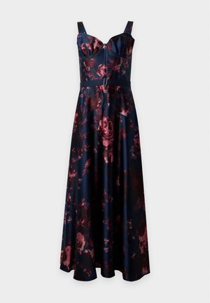 Femme portant une robe midi bleu foncé sans manches avec un imprimé floral rose et des escarpins noirs à bout pointu, debout devant un fond uni.
