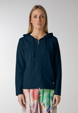 Hoodie navy cu fermoar, dotat cu glugă, două buzunare frontale și șnururi. Confecționat dintr-un material neted, asortat cu pantaloni colorate cu un model floral.