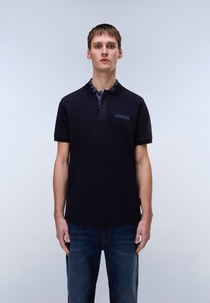 Young man wearing a navy polo shirt with logo patch on chest and dark blue jeans, standing against a plain light background.