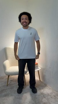 Mann med afro-frisyre står og smiler iført hvit t-skjorte og svarte bukser i et rom med teppe, en beige lenestol og et lite sidebord.