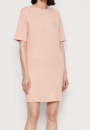 Kobieta ubrana w różową, sięgającą do kolan sukienkę typu T-shirt z krótkim rękawem i małym logo na górnej lewej stronie klatki piersiowej.