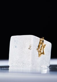 Cubetto di ghiaccio trasparente con una stella dorata incorporata, caratterizzato da una superficie liscia e piccole bolle d'aria, posizionato su una superficie riflettente.