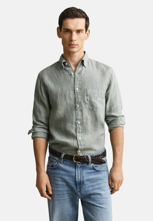 Mann trägt hellgraues Hemd mit aufgerollten Ärmeln, geflochtener dunkelbrauner Gürtel und hellblaue Jeans, steht vor weißem Hintergrund.