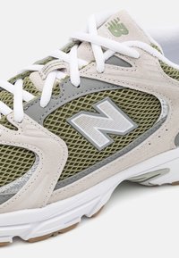 Chaussure de sport gris clair et vert olive avec tige en mesh, accents en daim, détail de logo et semelle en caoutchouc blanc avec motifs texturés.