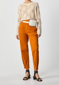 Beige pitsineulilla neulepaita, oranssit kapenevat housut, joissa on lahkeiden viimeistelyt, valkoinen vyölaukku ja mustat paksupohjaiset sandaalit, joissa on niittejä.