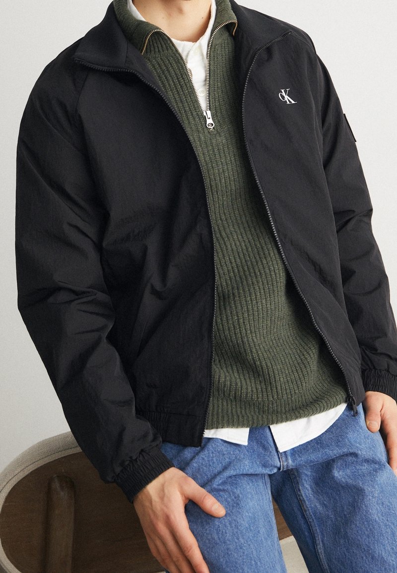 Veste noire zippée avec un pull côtelé vert en dessous, superposé sur une chemise blanche à col. Un jean bleu clair complète la tenue.