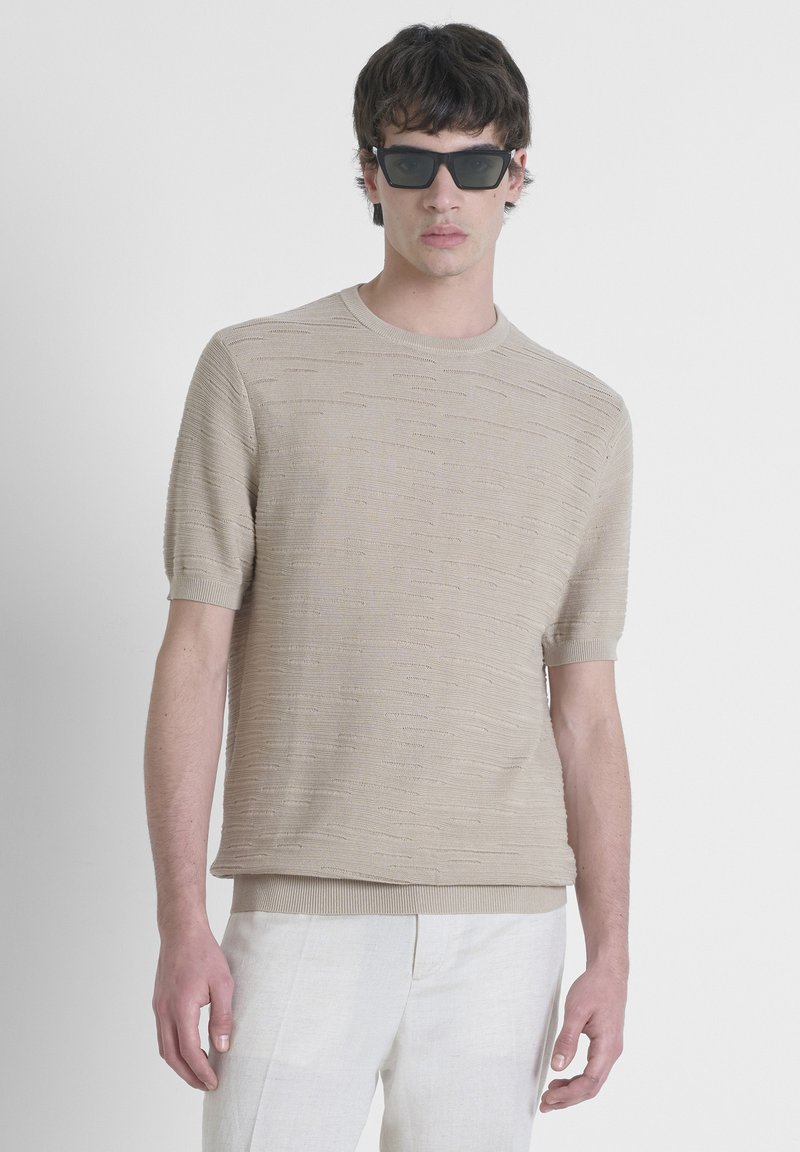Jersey de punto beige de manga corta con un patrón texturizado, escote redondo y dobladillo acanalado. Combinado con pantalones de lino claros y gafas de sol.