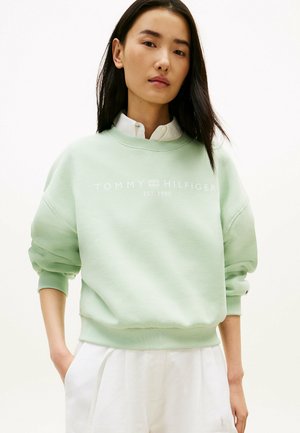Ung kvinde med langt mørkt hår iført en lysegrøn Tommy Hilfiger sweatshirt og hvide bukser, stående med den ene hånd i lommen.