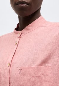 Közeli kép egy személyről, aki rózsaszín, gombos ingben van, melynek mellzsebén egy kis hímzett róka logó látható.