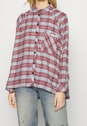 Žena v nadměrné červené a šedé kostkované košili s kapsou na hrudi, v kombinaci se světle modrými roztrhanými džínami.
