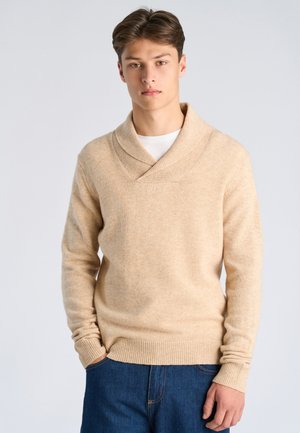 Joven con cabello castaño que lleva un suéter beige con cuello chal y jeans azules, de pie frente a un fondo claro y sencillo.