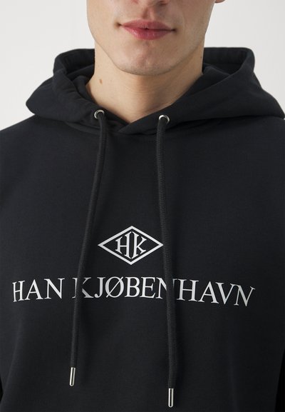 Črna kapuca s puloverjem iz mehkega materiala. Ima bel napis "HK" in "HAN KJØBENHAVN" natisnjen spredaj. Srebrni okrasni elementi.
