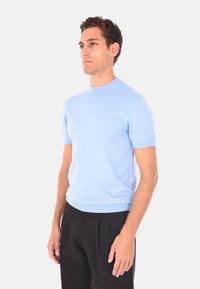 Uomo in piedi, con un maglione a maniche corte blu chiaro e pantaloni neri, di profilo a sinistra su uno sfondo grigio chiaro uniforme.