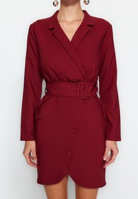 Burgundy omlottklänning i ett mjukt tyg, med långa ärmar, midjebälte, sidficka och knappar framtill.