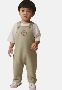 Gaismas zaļas kokvilnas kombinezons ar smaidiņa dizainu un koka pogām, kombinēts ar baltu T-kreklu un baltām slip-on kurpēm.