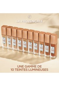 Dix flacons de fondation en verre carré avec des bouchons beige et marron. Les étiquettes affichent les noms des teintes et la marque. Les bases transparentes révèlent la couleur du produit.