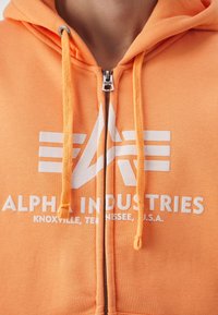 Felpa con zip arancione con una grande grafica bianca che presenta il testo "ALPHA INDUSTRIES" e un logo. Anche i cordini sono arancioni.