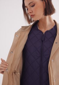 Giacca esterna color sabbia con bottoni a pressione e zip, sopra uno strato interno in nylon trapuntato blu scuro con collo tondo e texture a contrasto.