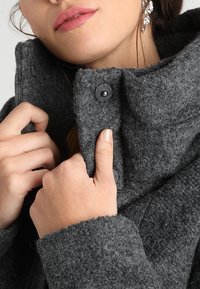 Grijze wollen jas met een hoge kraag, gestructureerde stof en zwarte knopen. Close-up die een hand toont die de rand van de kraag vasthoudt voor warmte.