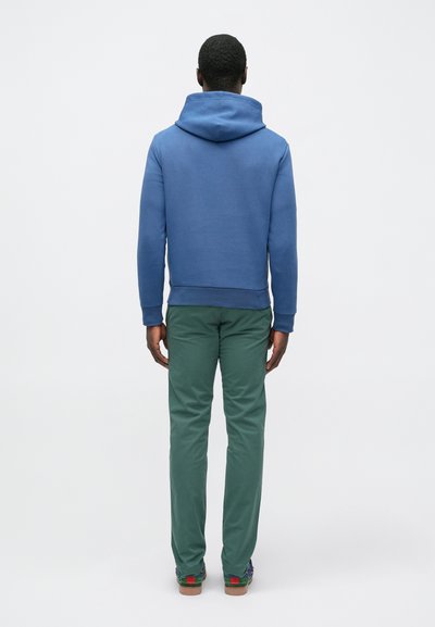 Άνδρας που στέκεται με την πλάτη, φοράει μπλε φούτερ με κουκούλα, πράσινο παντελόνι και πολύχρωμα αθλητικά παπούτσια, μπροστά σε απλό φωτεινό φόντο.