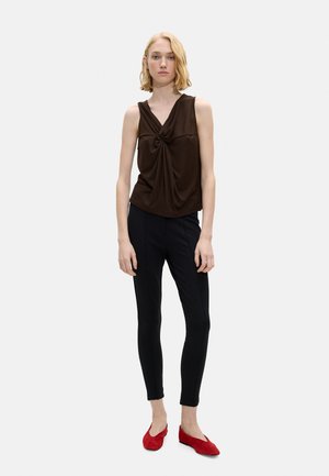 Młoda kobieta z krótkimi blond włosami w ciemnobrązowym bezrękawniku z efektownym skręceniem, dopasowanych czarnych spodniach i czerwonych płaskich butach, stojąca na białym tle.