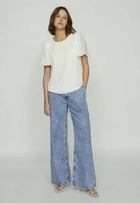 Kvinne som har på seg en hvit kortermet bluse med pufferm, lyseblå vide jeans og hvite hælsandaler, stående med den ene hånden i lomma.