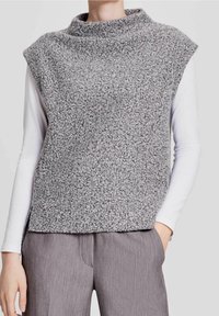 Person som bär en texturerad grå ärmlös tröja över en vit långärmad skjorta och grå byxor, med ena handen i fickan.