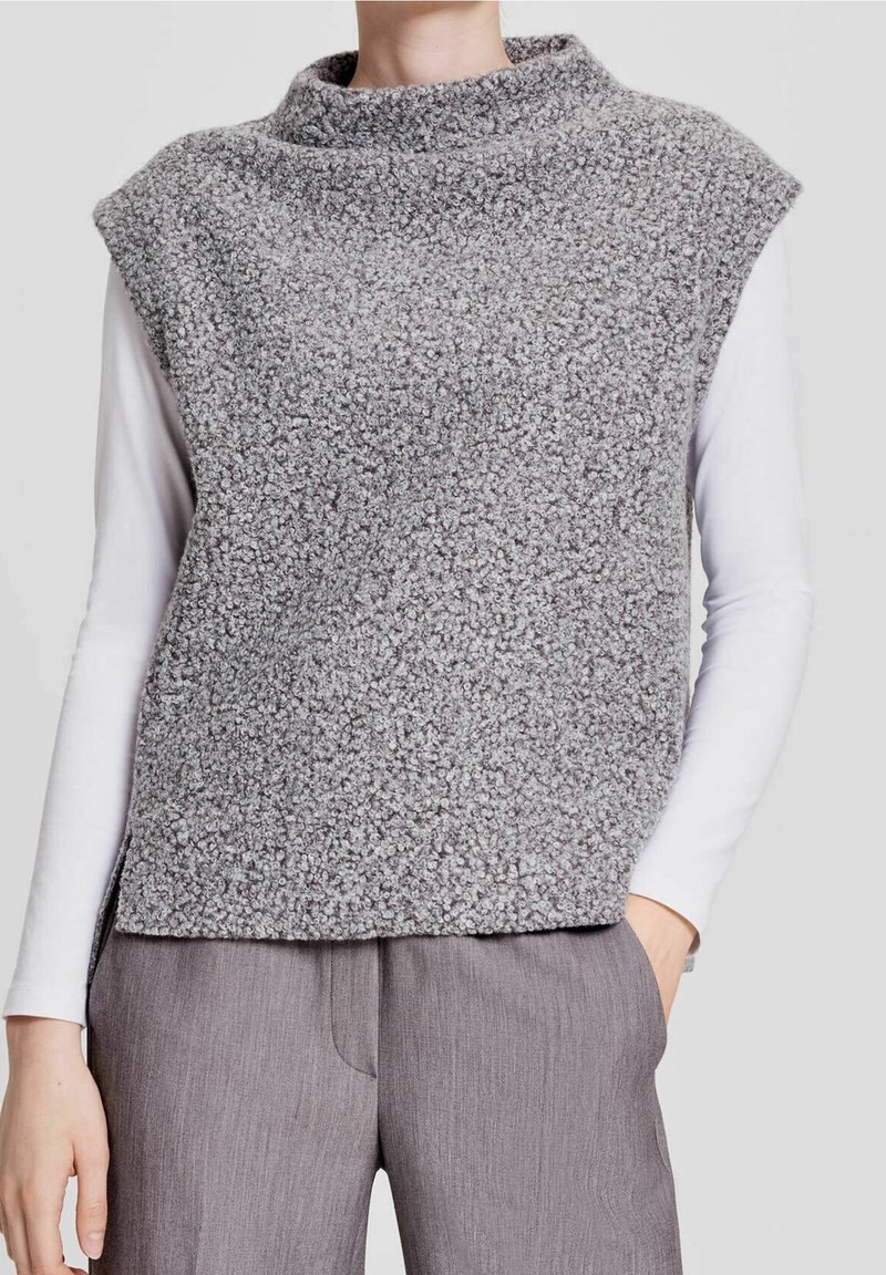 Person som bär en texturerad grå ärmlös tröja över en vit långärmad skjorta och grå byxor, med ena handen i fickan.