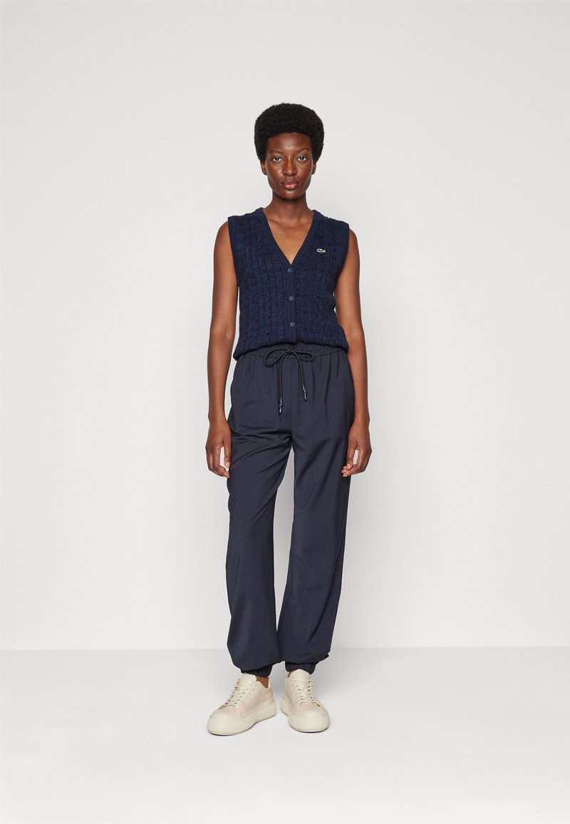 Granatowy sweter bez rękawów z dekoltem w serek oraz guzikiem z przodu, w połączeniu z luźnymi granatowymi spodniami i beżowymi sznurowanymi trampkami, stojąc na jednolitym tle.