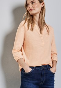 Blusa de color melocotón con escote en V y tres botones decorativos. Presenta mangas recogidas y una textura suave y ligera. Combinada con jeans oscuros.